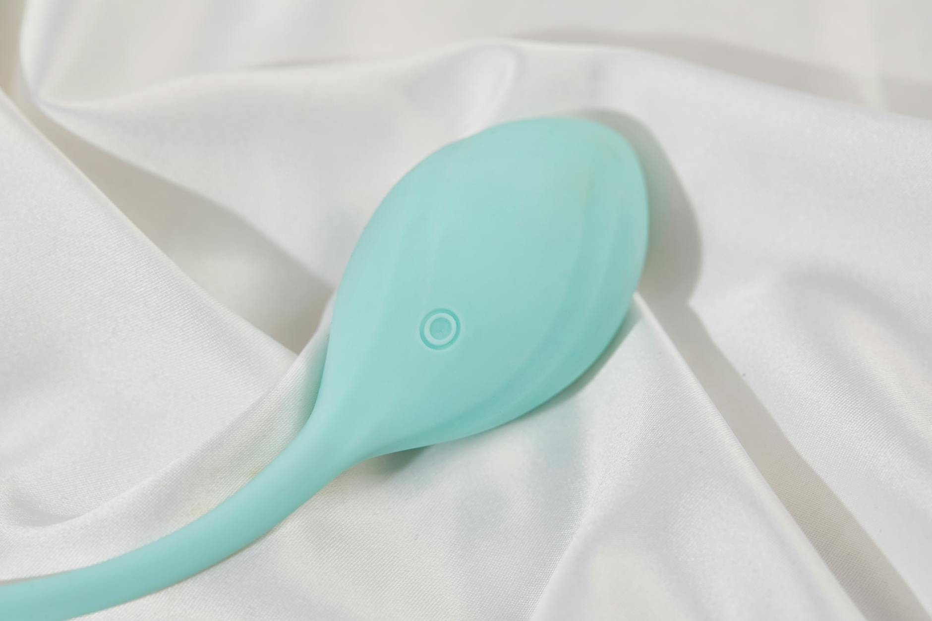A teal clitoral vibrator resting on smooth white silk fabric