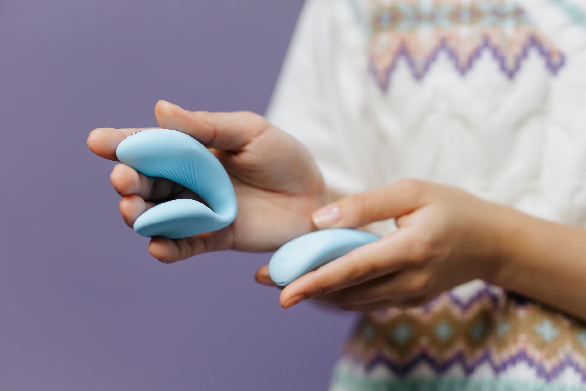 Two people's hands together holding a blue vibrator against a purple background, representing shared pleasure exploration.